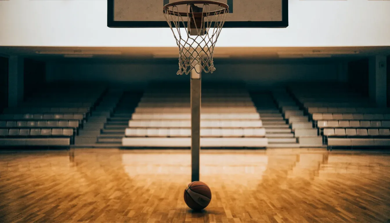 Ein Basketballkorb hängt in einer leeren Halle über einem Holzparkett, von der Decke fällt gleichmässiges Licht auf den Boden.