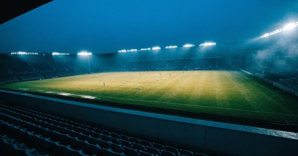 Ein Fussballspiel am Abend aus der Tribünenperspektive, die Spieler sind nur als kleine Figuren zu erkennen und das Flutlicht beleuchtet den nassen Rasen.
