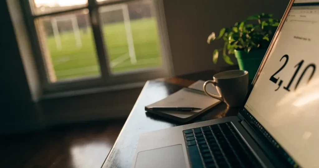 Ein aufgeräumter Schreibtisch mit Pflanze und Tasse in einem Heimbüro, vor dem Fenster liegt ein Fussballplatz im Gegenlicht.