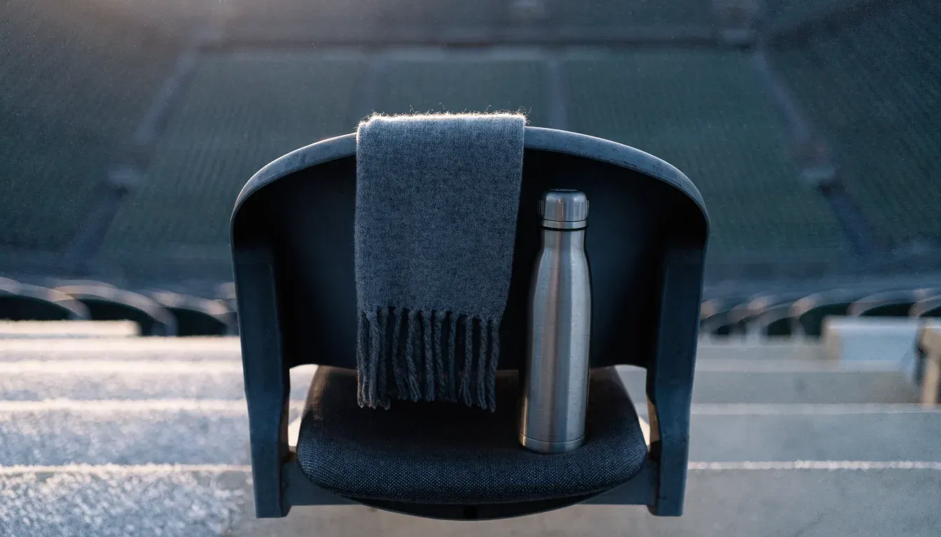 Ein grauer Wollschal und eine Thermoskanne liegen auf einem Sitz im winterlichen Stadion, Raureif liegt auf den Stufen.